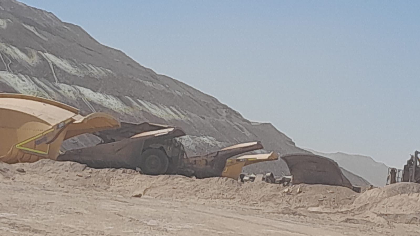 Several large industrial dump trucks positioned on a dusty construction site with a mountain in the background.